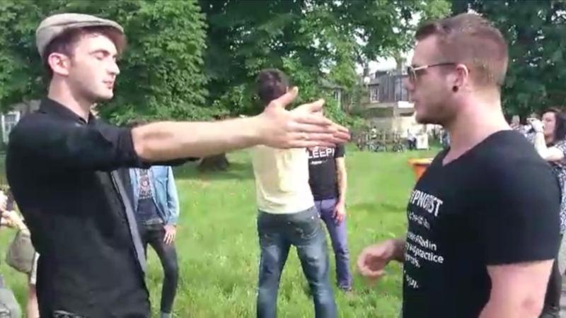 Rory Z Fulcher performing street hypnosis, about to use the magnetic hands induction with a young man at a fair, with people milling around in the background.